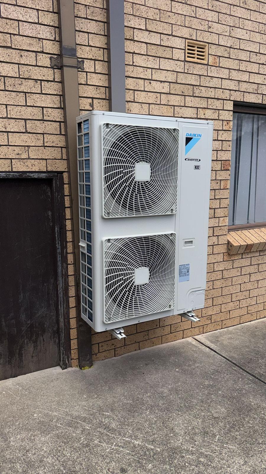 Large twin-fan Daikin Inverter outdoor unit mounted on cream brick wall